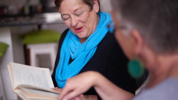A woman with short hair and bright blue scarf enthusiastically discusses a book with another another woman at a library book club.