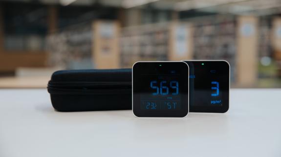 Two air quality monitors on a table in front of a library, displaying different air quality readings against a backdrop of bookshelves.