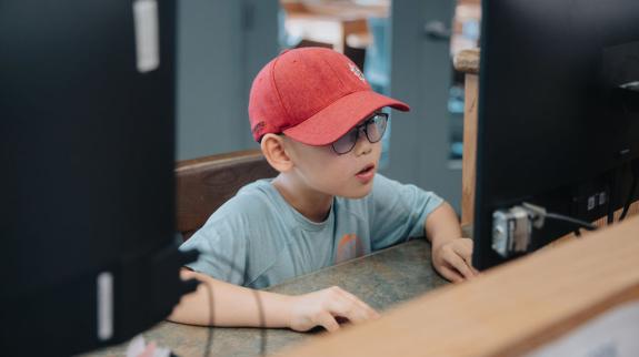  A young boy in a red hat and sunglasses sits at a computer, focused on the screen in front of him.