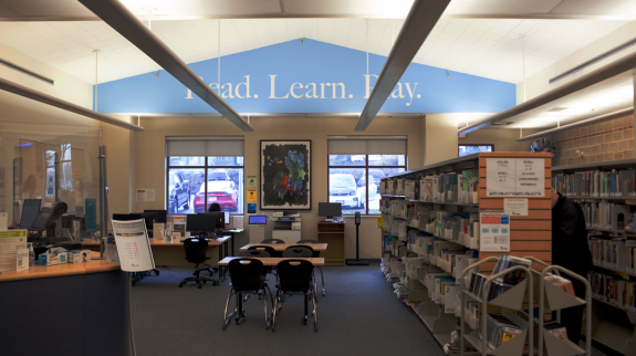Interior of Dean Drysdale Library with feature wall that reads "Read Learn Play" above rows of bookshelves.