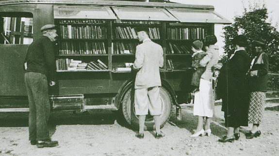 People of various ages stand around a vibrant bookmobile truck, discussing books and enjoying the community atmosphere.