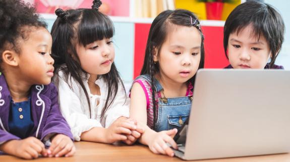 Children gathered around a table, engaged with a laptop, exploring stories on Bookflix together.