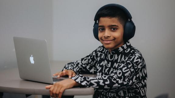 A boy works on a laptop computer in the library. He is wearing headphones.