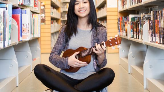 A young woman sits on the floor, strumming a ukulele with a joyful expression on her face.