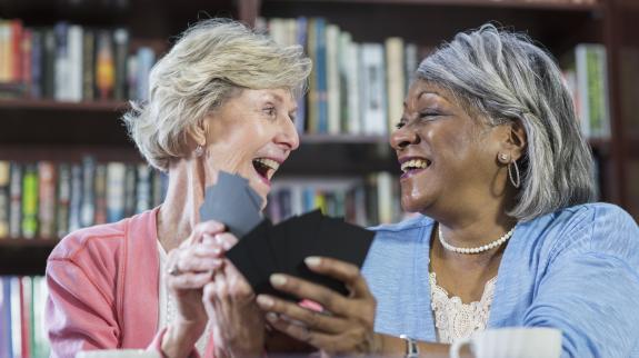 Two older women joyfully laughing together while each holding a hand of playing cards, sharing a moment of connection and friendship.