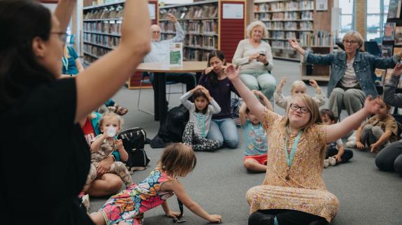 A lively scene in a library where children are seated, all raising their hands enthusiastically as they participate in Storytime.