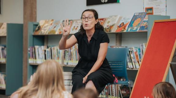 A woman in a black shirt sits while telling a story, holding up four fingers, in front of a group of attentive children. A empty red felt board sits to her right.