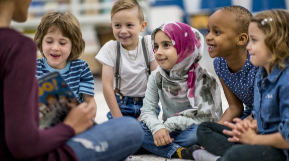 A diverse group of four children sitting on the floor. An adult reads happily to them in a cozy setting.