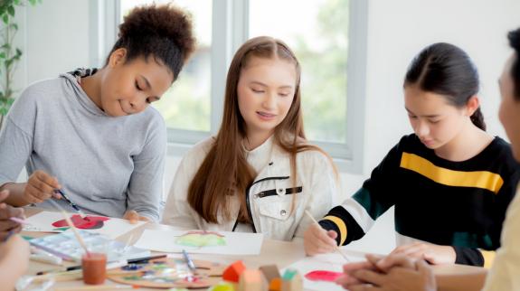 A group of 3 diverse teen girls sitting around a table colouring, drawing, and painting. 