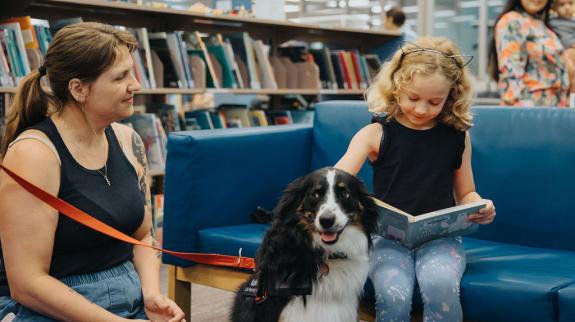 A young girl sits on a blue couch petting and reading to a black and white dog. The dog an a caregiver sit on the floor next to the reading child.
