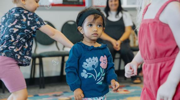 Three toddlers try to catch bubbles during Toddler Social, while a parent watches in the background.