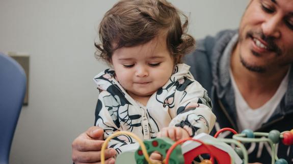 A smiling dad and a baby girl engage in playtime, surrounded by a variety of fun toys at the library.