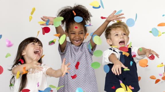 A multi-ethnic group of three small kids, throw colourful confetti into the air while laughing.