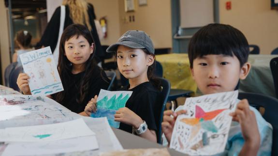 Asian boy (8) and two Asian girls (8) hold up their painted pictures.