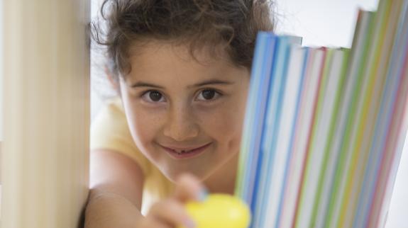 A young female child peeks through a bookshelf to grab a yellow Easter egg.