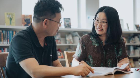A man and a woman are sitting together at Maple Ridge Public Library. An open book sits on the table.