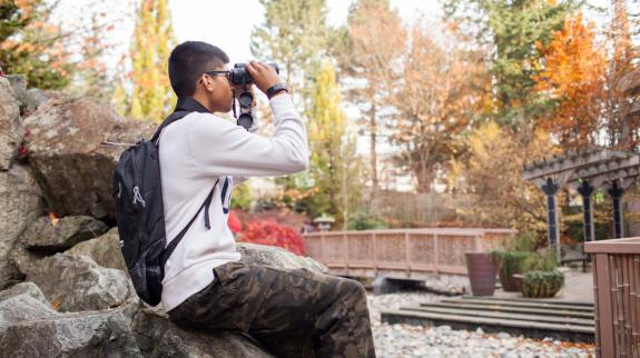 South asian teen boy (18) uses with binoculars and backpack looks at birds in the distance.