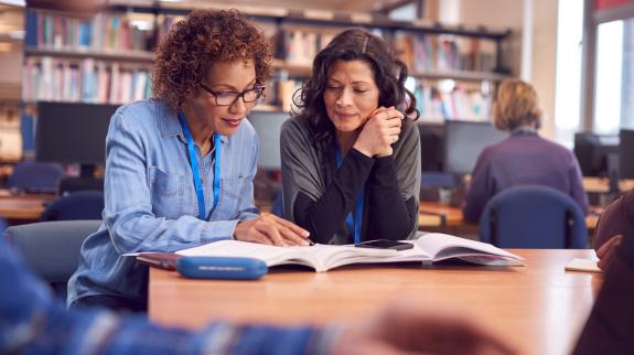 Two women sit together at table in library discussing and working on their writing skills. Library stacks, and public computers are used by other customers in the background.