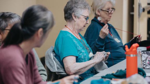 Four older ladies sit in a row at a table. They are all working on different knitting and crocheting projects and enjoying each other's company.