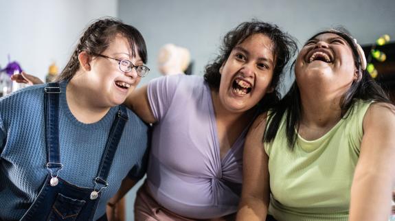 Three young female adults with diverse abilities embrace and laugh with each other.