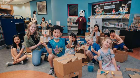 A large group of children and a female library staffer, cut up cardboard using Makedo tools and screws to create imaginative cardboard creations like a dragon head.