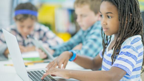 A multi-ethnic group of children are indoors in a library program. They are wearing casual clothing. They are working together on a laptop computer learning to code together.