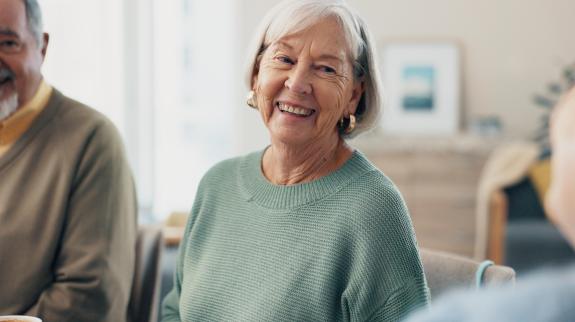 A group of seniors gather to socialize. A senior woman looks into the camera and smiles, while a man appears in the side of the image.