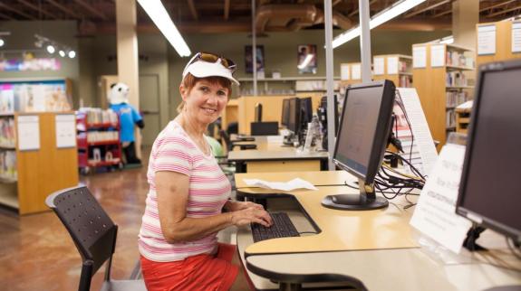 A older woman  in shorts, t-shirt, and hat, sits at a library computer in a library. She looks at the camera while typing. Bookshelves can be seen in the background.