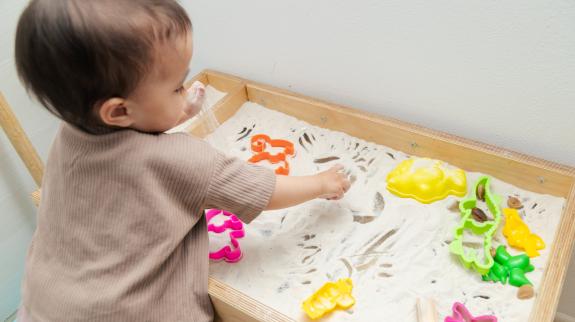 Toddler playing kinetic sand with plastic shapes on sensory table.