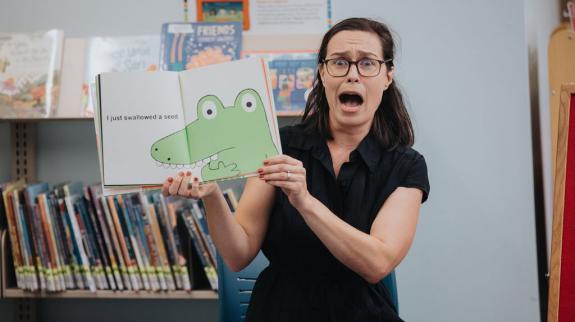 A librarian reads a storybook to a group of kids. Her face makes an animated scared expression as she shows the kids the image of a crocodile in the book.