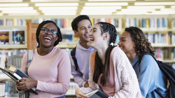 A multiracial group of four high school students carrying backpacks, laptops and textbooks, walking in a library, conversing, laughing. They are teenagers, 15 and 16 years old. 