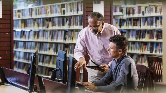 Two multi-ethnic men in the library looking at a computer screen and conversing. A mature man in his 40s is sitting at the desktop PC and a senior African-American man in his 60s is standing beside him.
