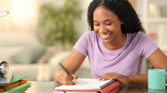 Teen girl, sitting at a counter writing on paper that is stacked on top of a binder. A coffee mug and other desktop items can be seen in the surrounding area.