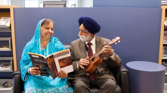 South Asian man (70s) strums a ukulele while a South Asian woman (70s) holds a music book open for him.