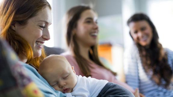 Smiling mother holding sleeping newborn son sitting by friends at a mom's group. 