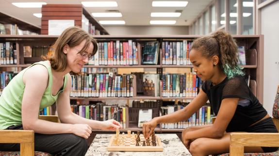 Caucasian woman (20s) and African-American girl (12) play chess on a low white table, surrounded by library stacks. 