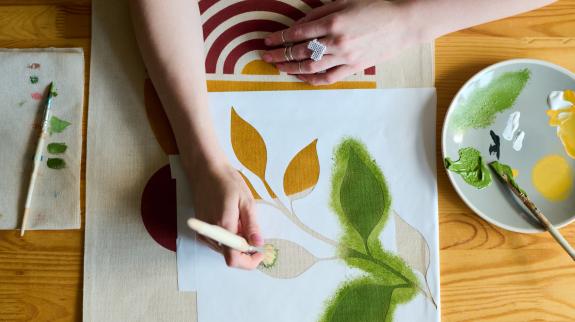 Above angle of hands of young female artist with paintbrush painting green leaves on paper while sitting by table in workshop.