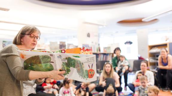 Female library staffer (50s) reads 'The Very Hungry Caterpillar' to a large audience of kids and parents. Audience sits on a blue carpet on the library's floor. 