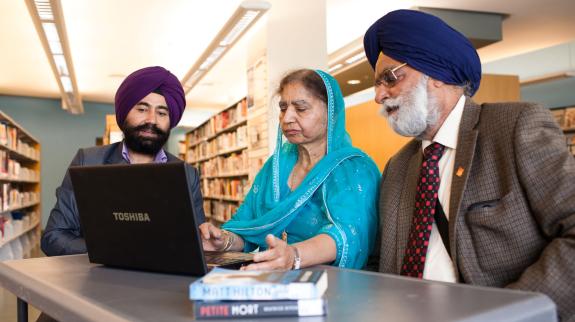 A South Asian man with purple turban assists a South Asian woman wearing a teal langa and her South Asian man in blue turban with a laptop.