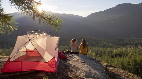 Young women watch the sunset on a mountain ledge campsite. Their tent is illuminated in the sunlight.
