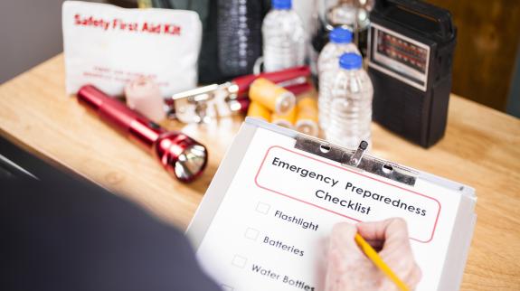 Unrecognizable woman holds emergency preparedness checklist on clipboard with pencil and prepares a large pile of supplies to be used in case of a natural disaster including: flashlight, batteries, water bottles, first aid kit, lantern, radio, backpack, can opener, mask. Over the shoulder view.