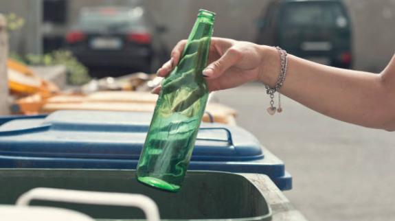 Women's hand putting an empty glass bottle into a green recycling bin