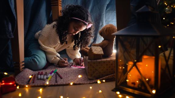 Little girl writing down letter under the table surrounded by fairy lights spread out over the floor.