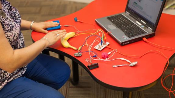 A person plays with a Makey Makey circuit board device by connecting it to various items to create computer commands.