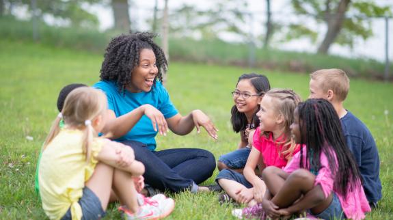 A female librarian with a group of children in the park. She is telling them a story. The children are happy and smiling.