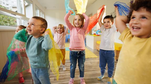 Group of happy children in playroom dance with scarves in their hands.