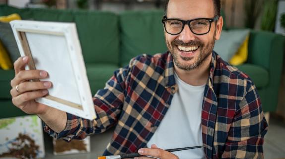 A smiling man in a casual plaid shirt enjoys the moment while painting on a canvas.