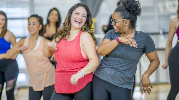 A multi-ethnic group of adult women are dancing in a fitness studio. They are wearing athletic clothes.