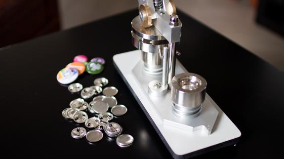 A button making machine sits on a table with pins and covers sitting nearby.