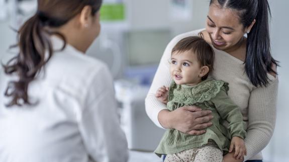 A young mother brings in her little toddler for a check up. She has the girl seated on her lap as she talks with the doctor about her health.